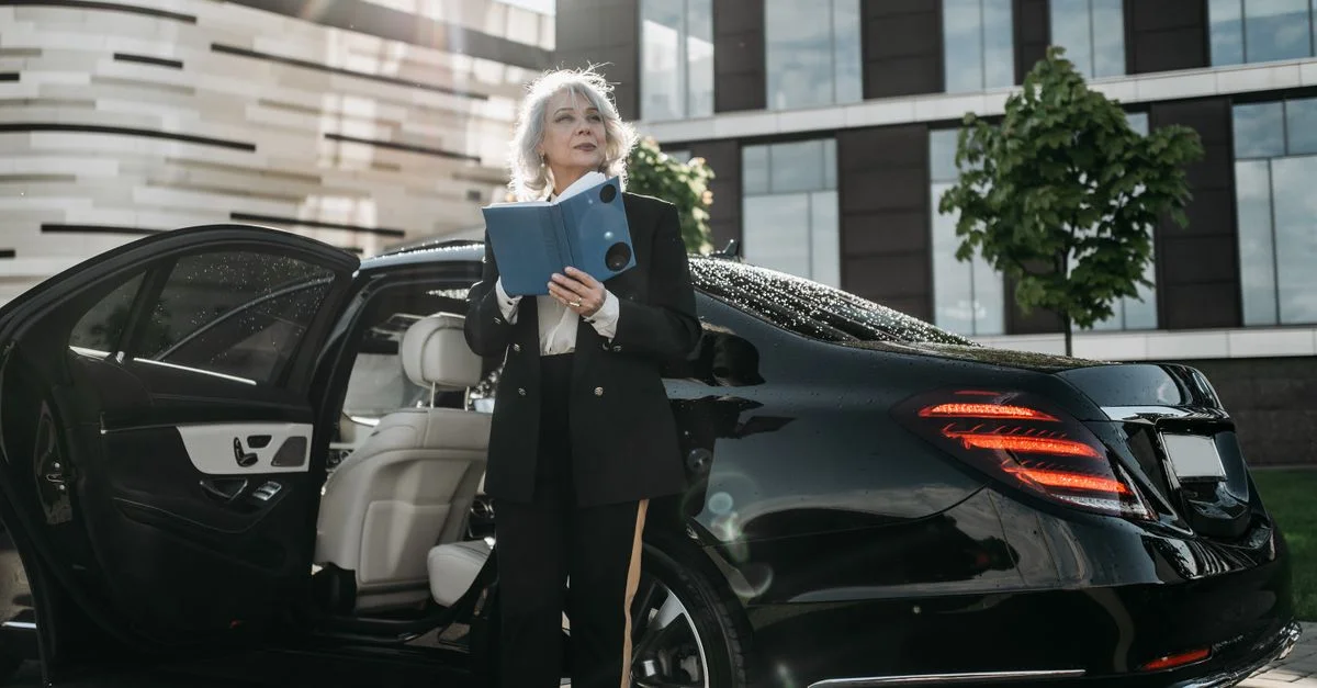 Une femme d'affaires senior sort d'une voiture de luxe noire, tenant des documents près d'un immeuble de bureaux moderne.